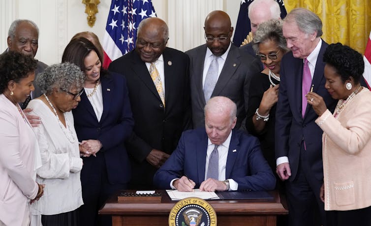 A an at a desk signing a piece of paper with many people standing around him, watching.